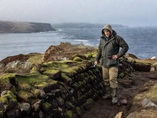 Researcher standing beside sod-covered turf wall foundations overlooking a cold North Atlantic bay under natural light
