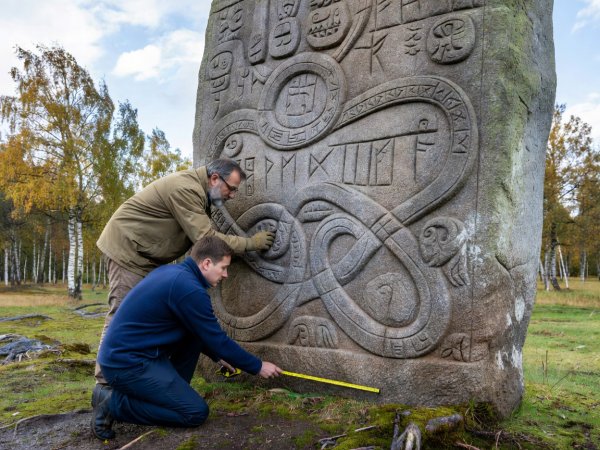 Epigrapher in natural light examining deep runic grooves and ribbon ornament on a granite monument in Scandinavia